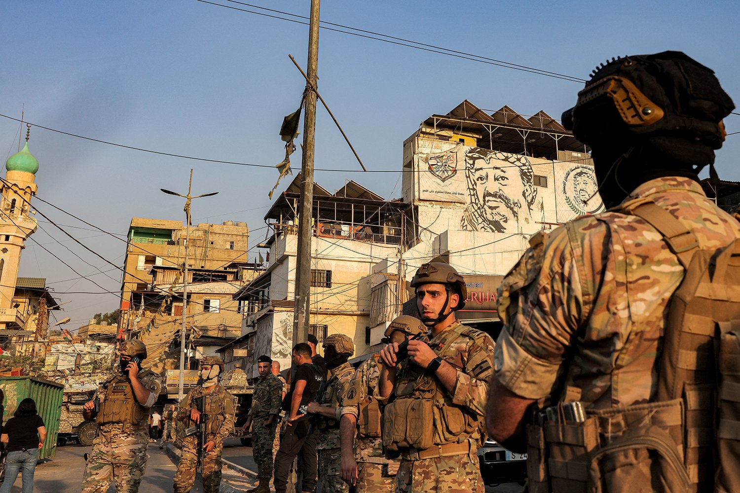 Soldiers in uniforms and helmets in front of a refugee camp. A depiction of Yasser Arafat is seen on the edifice of a building in the distance.