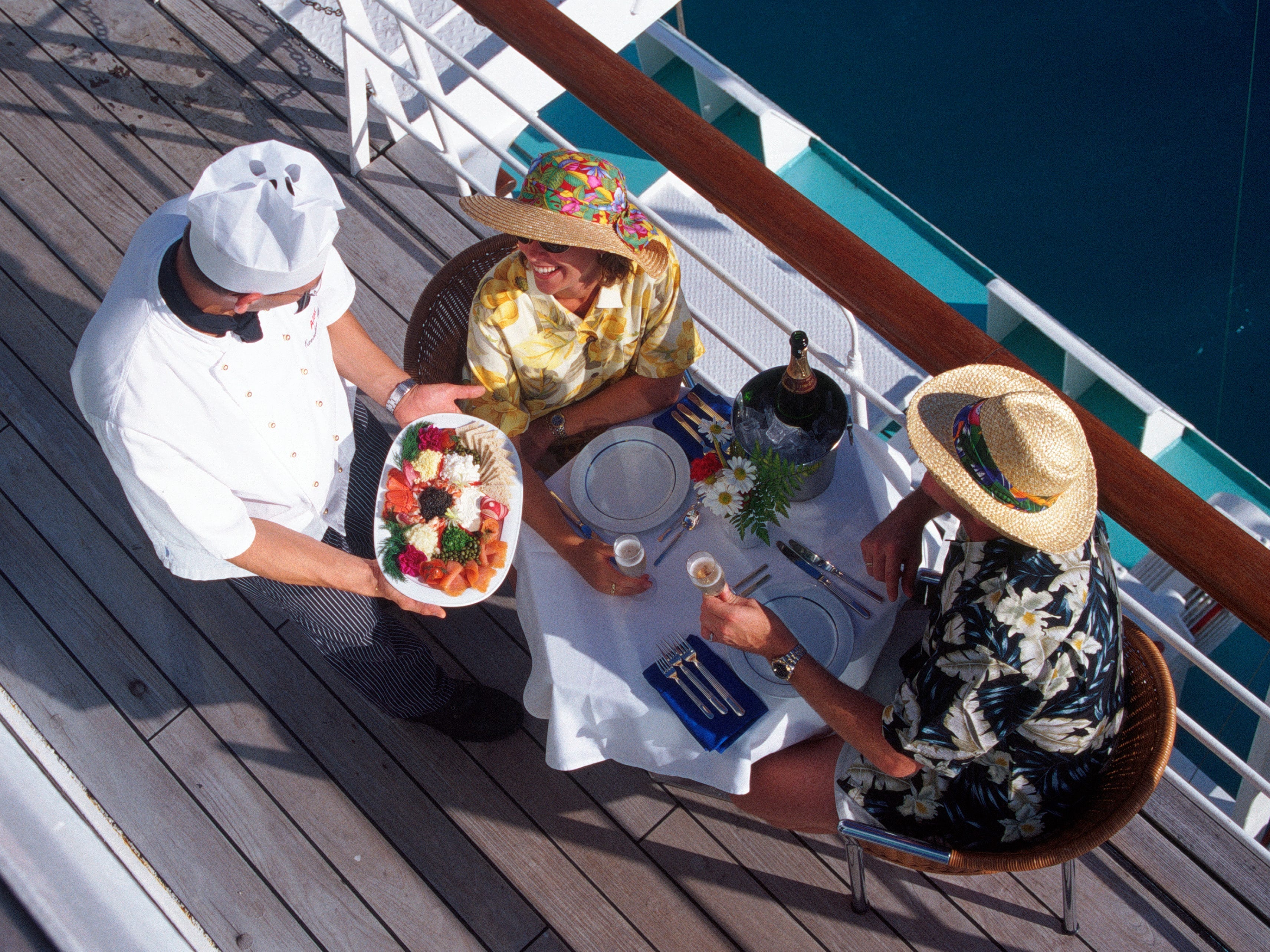 A waiter serving food to a couple sitting at a table on the balcony of a cruise ship.