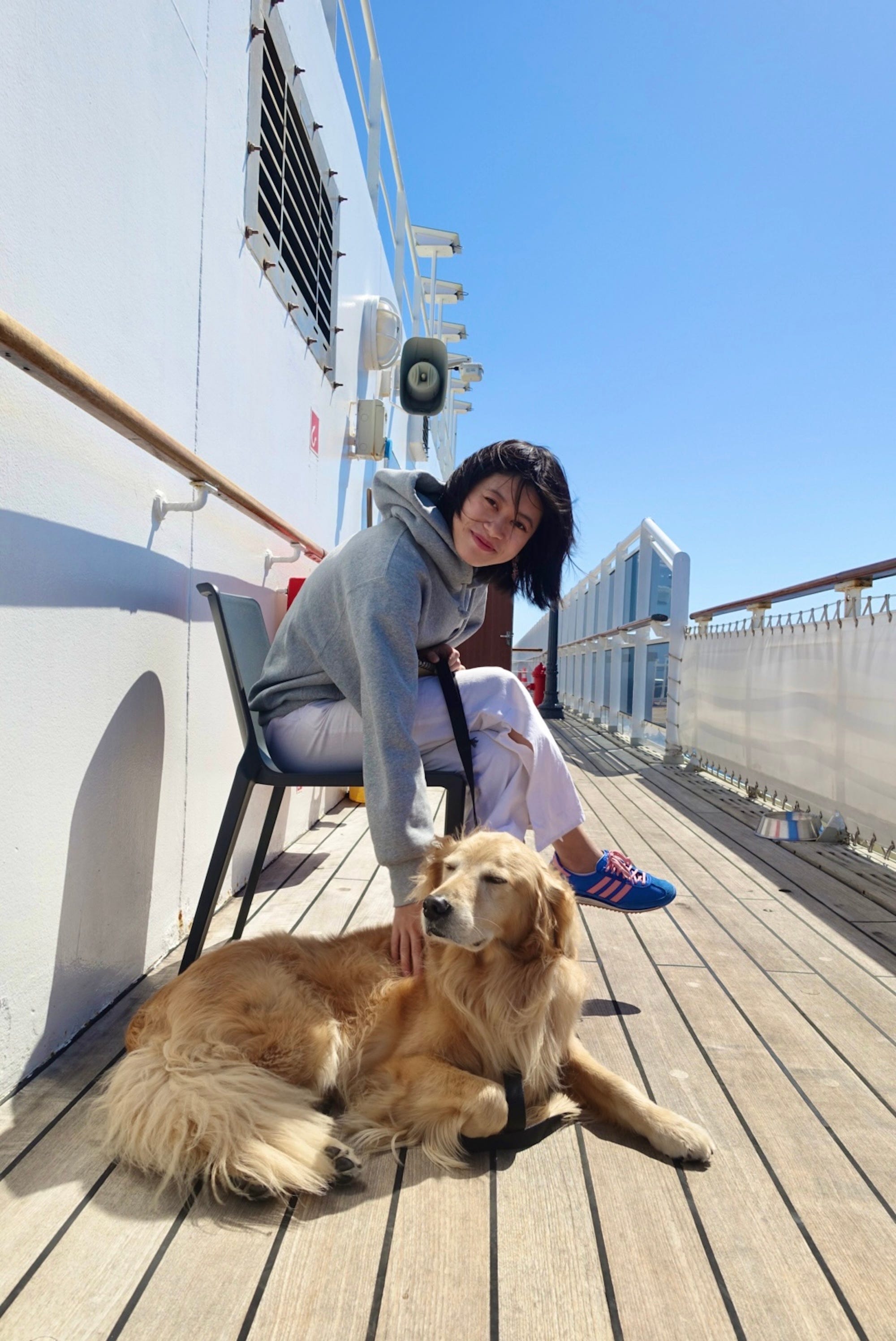 A woman leans down from a chair to pet her dog on the deck of a cruise.
