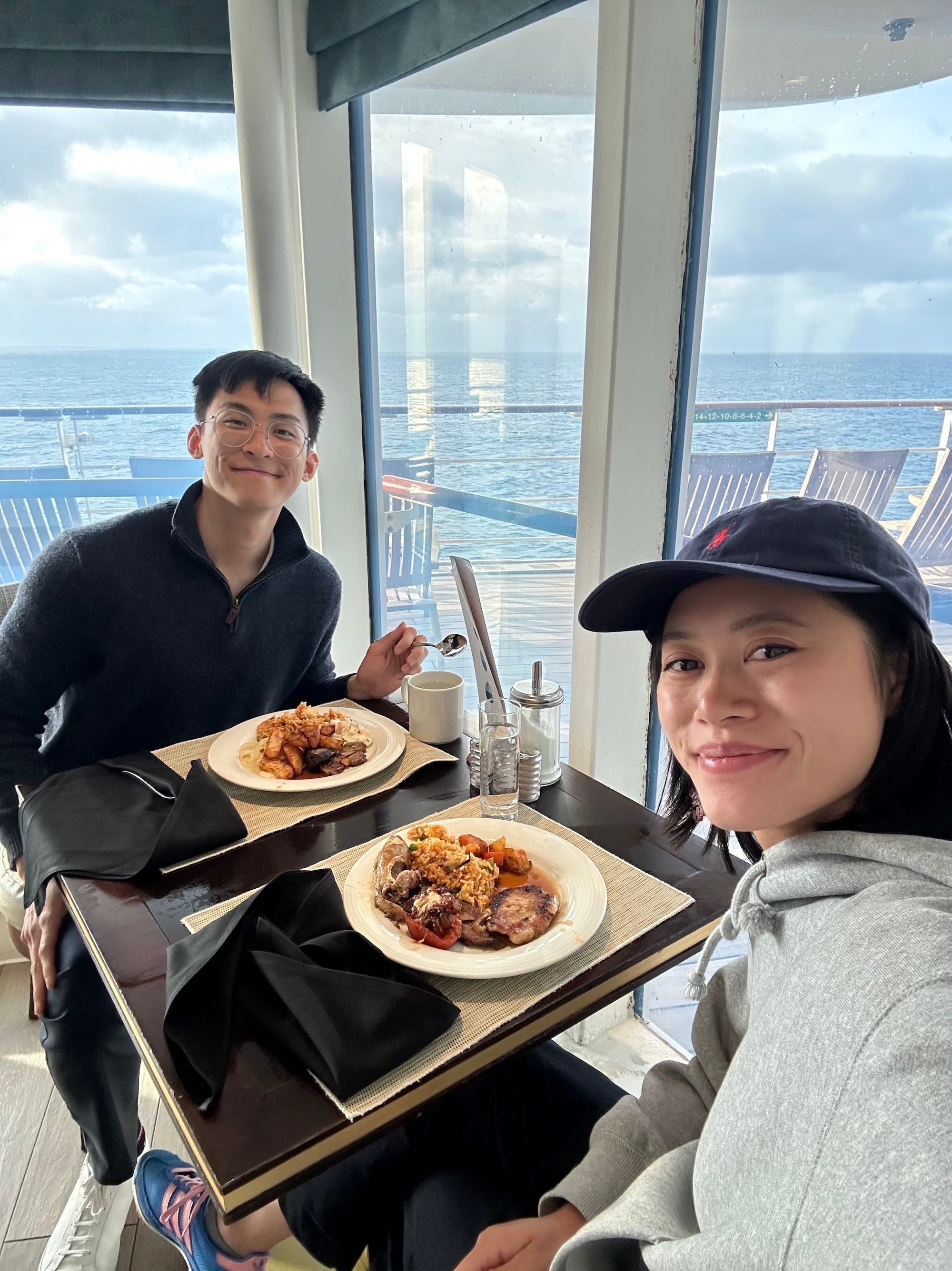 A couple smiles in a selfie as they eat dinner on a cruise. The deck and ocean are visible through a glass wall.