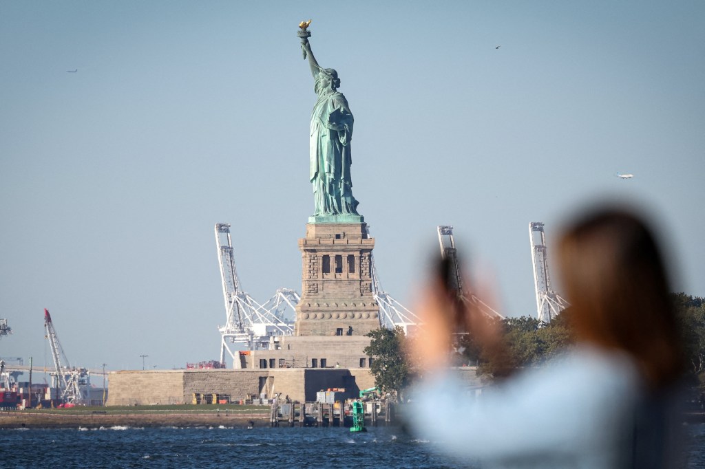 A blurred person in the foreground taking a photo of the Statue of Liberty, with the base of the statue and large cranes visible in the background.