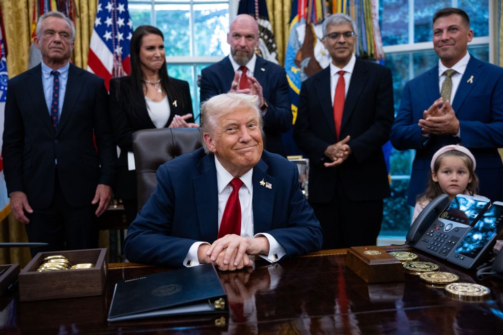 President Trump smiles while seated at a desk, surrounded by several individuals, including a small child and Robert F. Kennedy Jr., in the Oval Office.