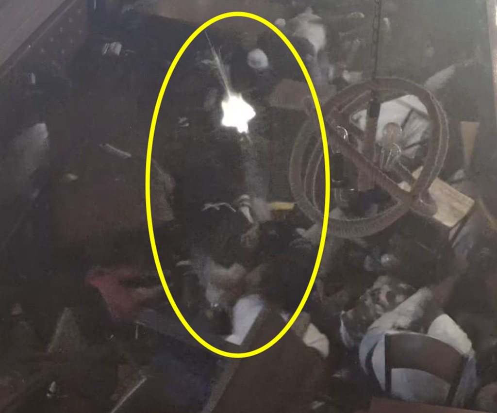 Still image from security footage showing a muzzle flash from a gun fired inside a dark room.