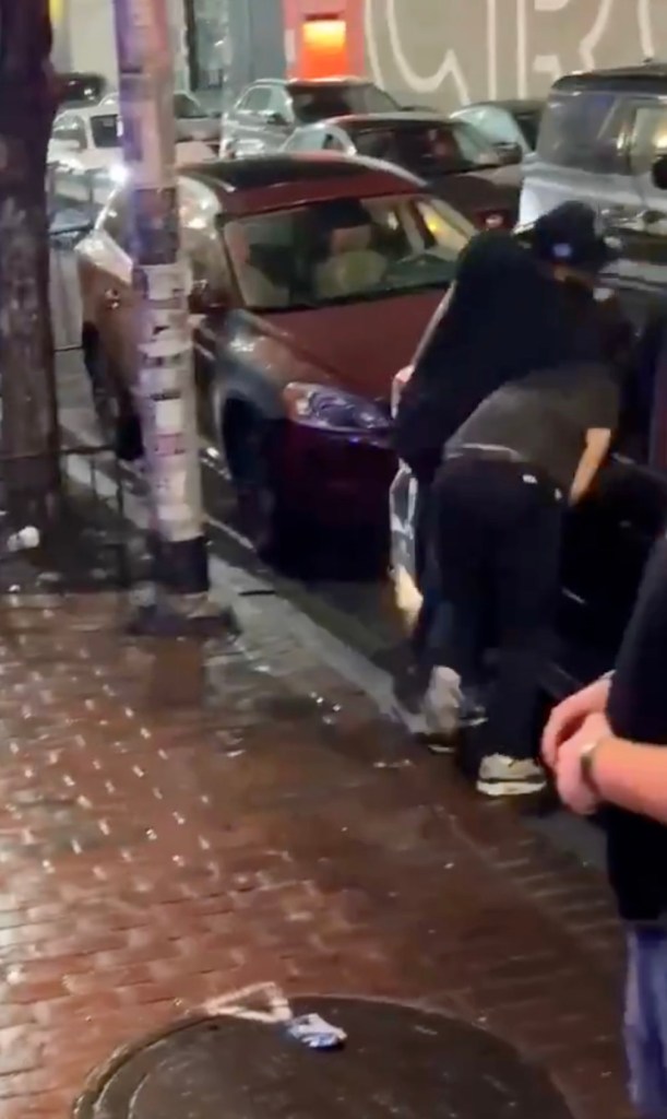 Two men grappling near a car on a city street at night.