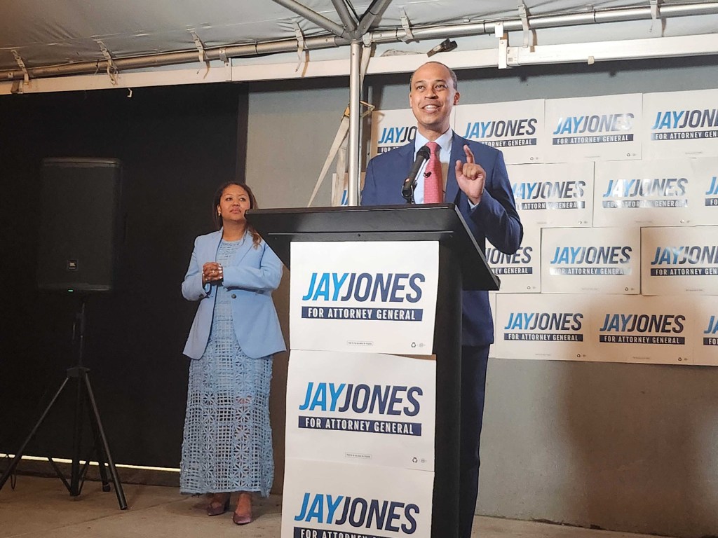 Jay Jones speaking from a podium with his wife, Mavis Jones, standing to his left, in front of a backdrop of campaign signs.