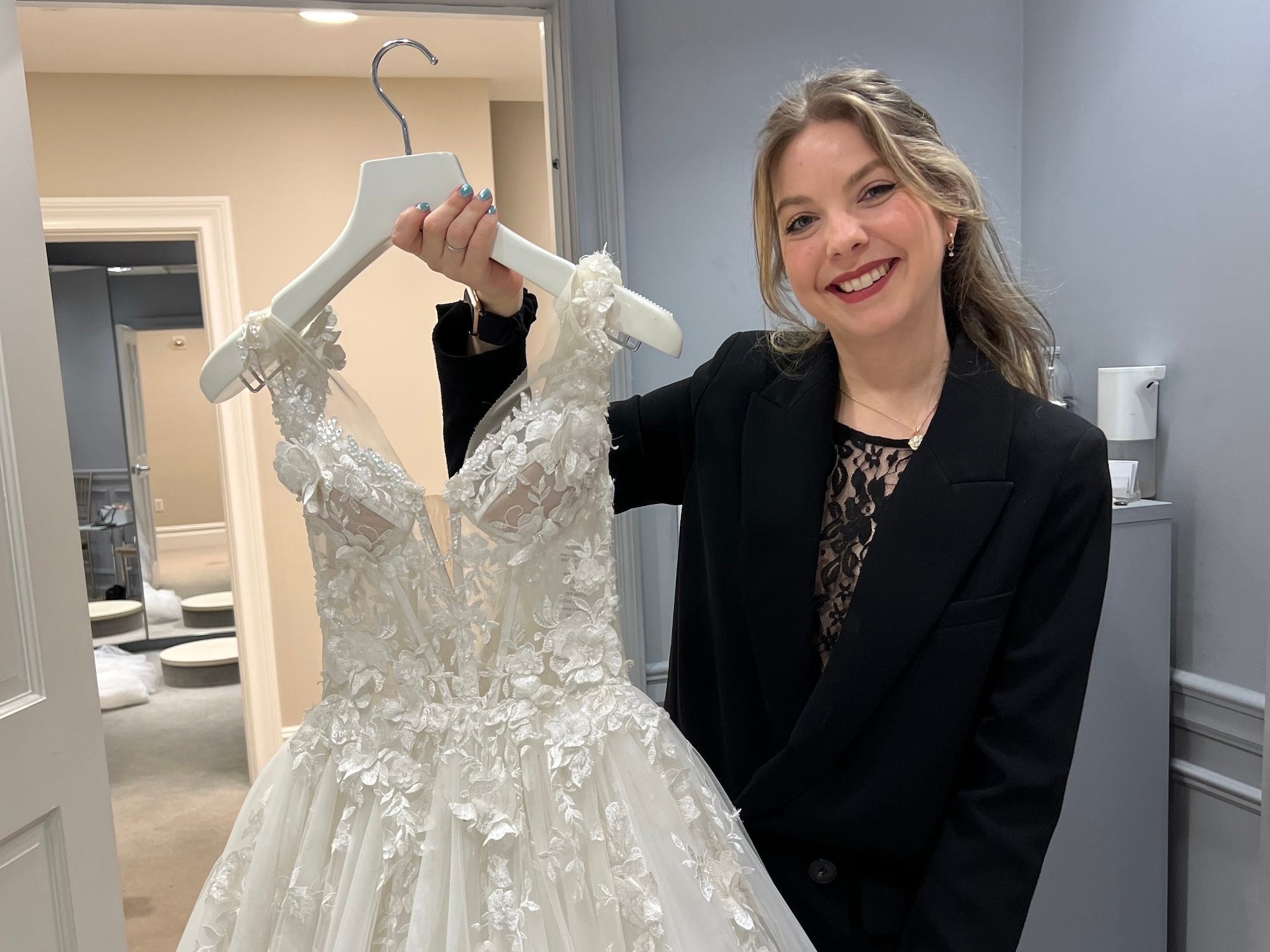 A woman in a black outfit holds up a wedding dress on a hanger in a blue room.