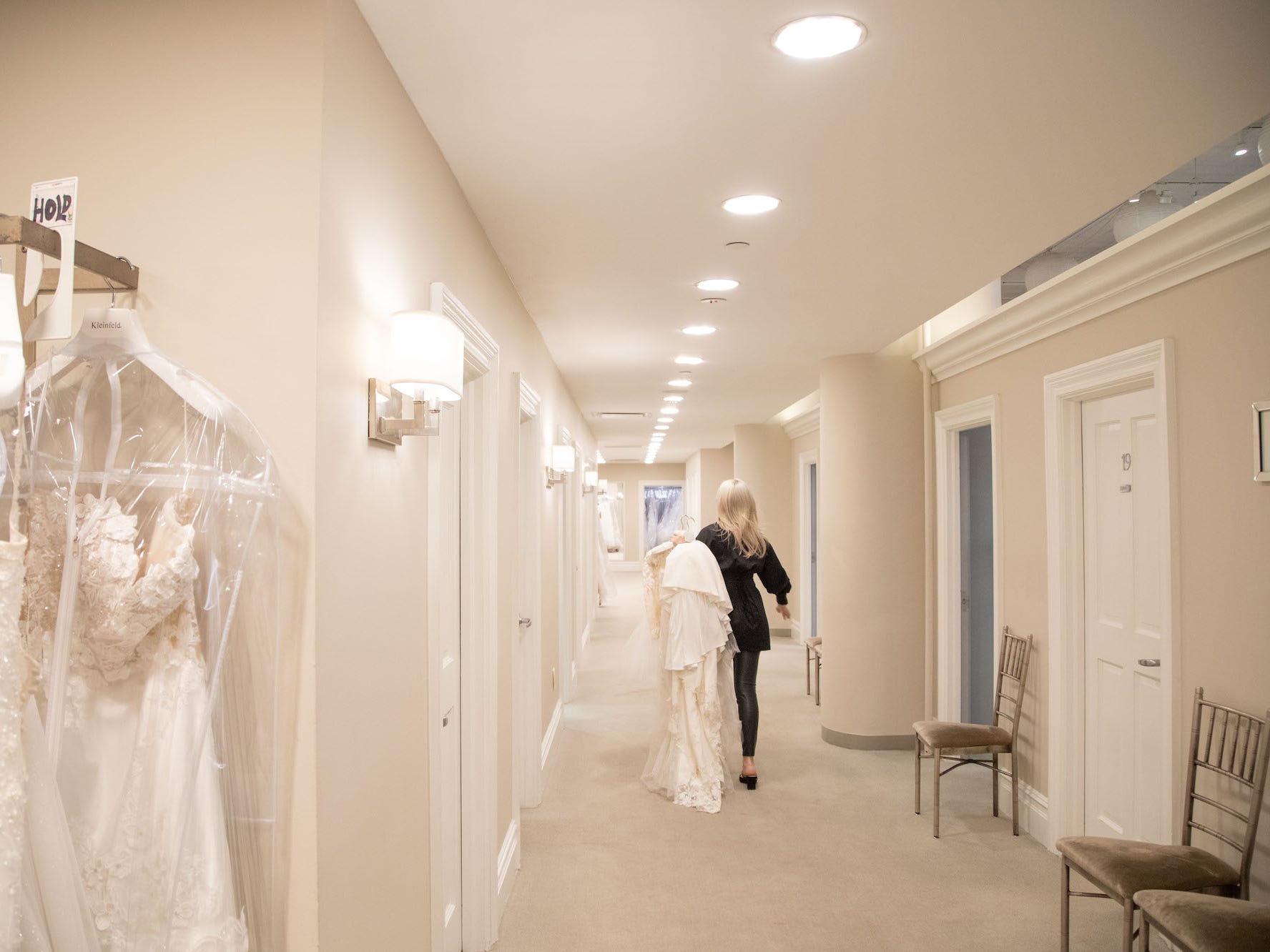 A woman walks down a hallway with doors carrying a wedding dress.