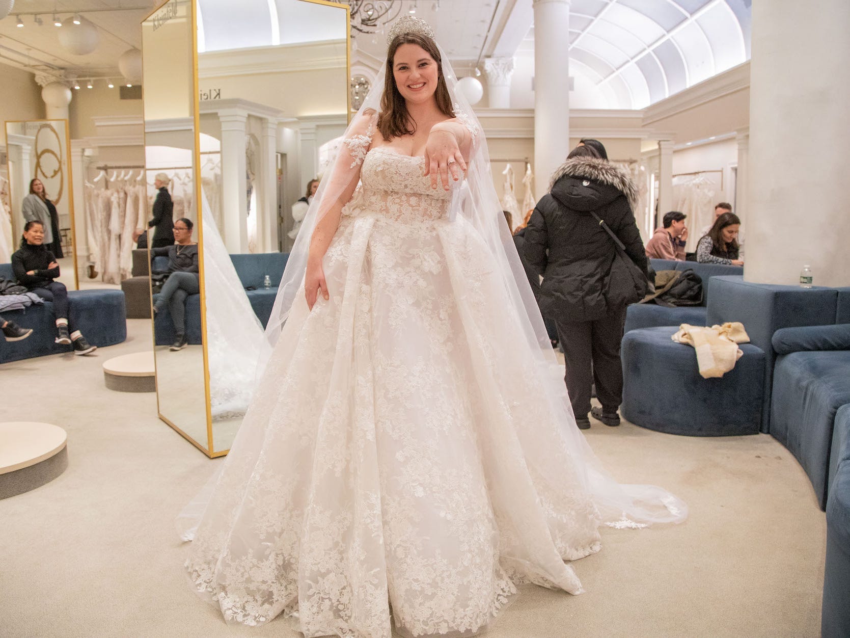 A woman holds her hand out with an engagement ring on it as she poses in a wedding dress.