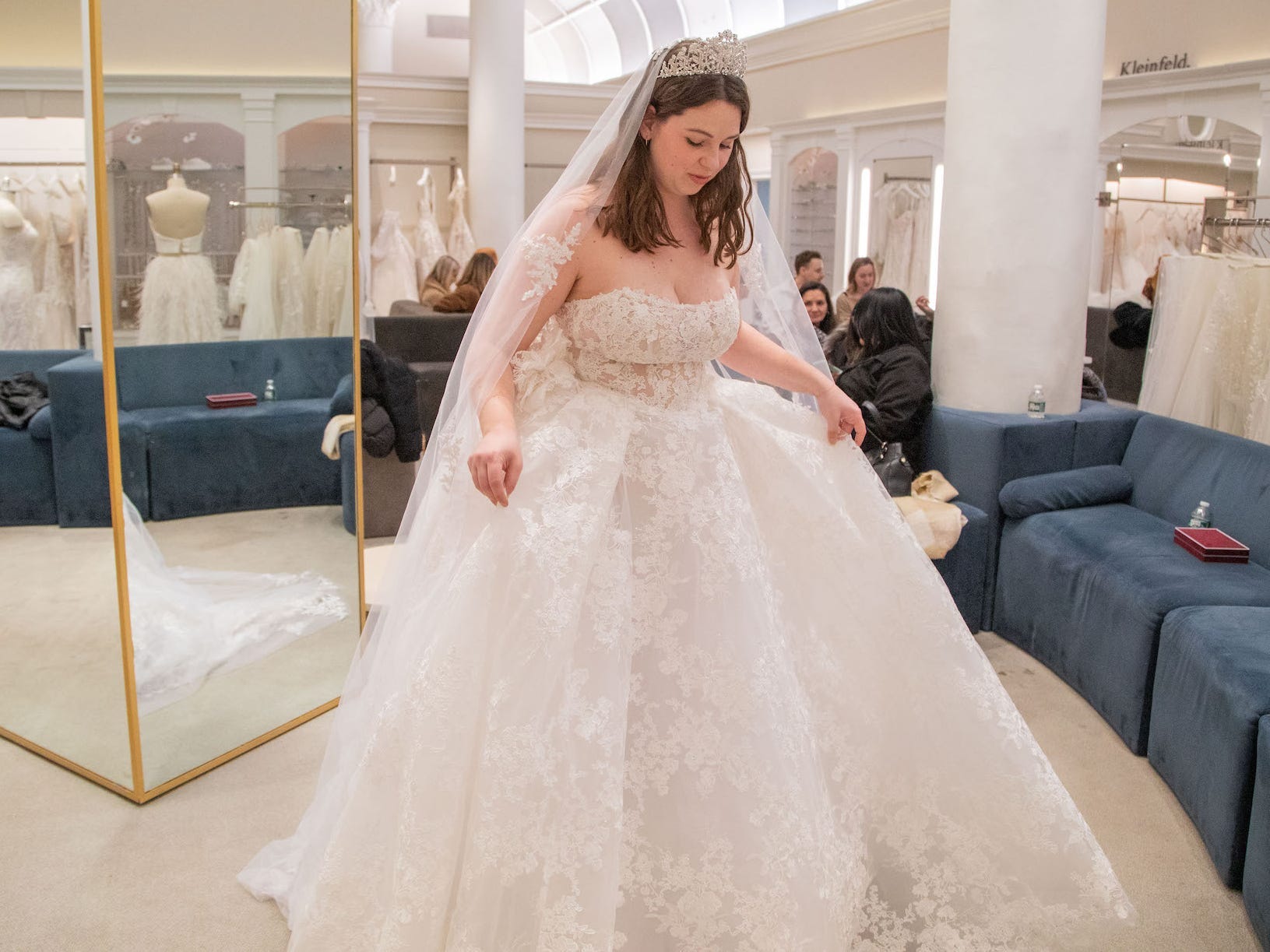 A woman looks down at the skirt of a wedding dress she's wearing in a bridal boutique.