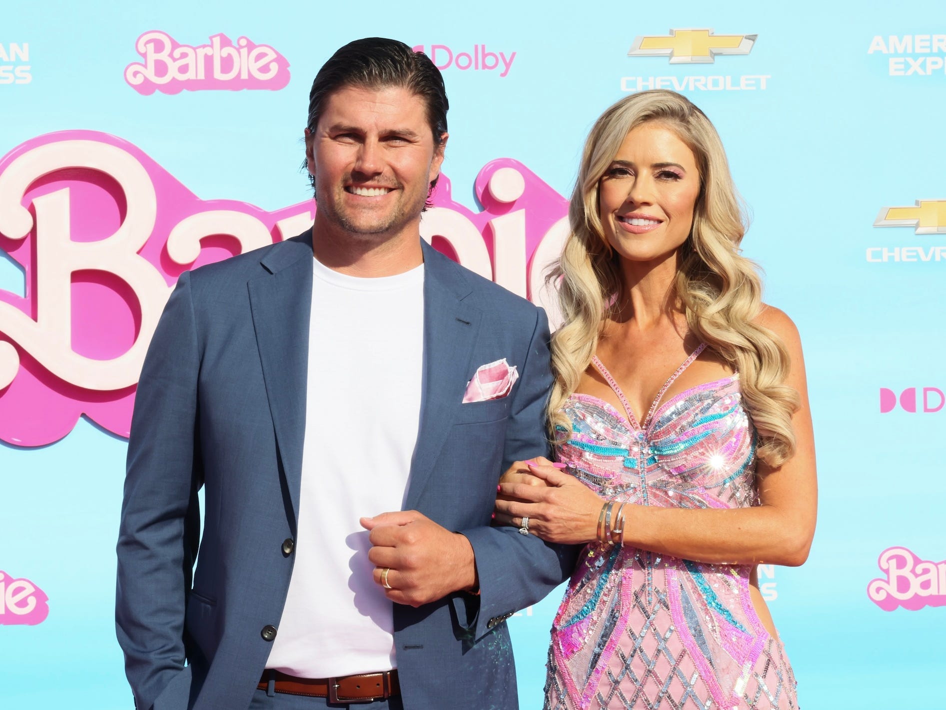 Josh and Christina Hall pose in front of a blue backdrop with the Barbie logo on it.