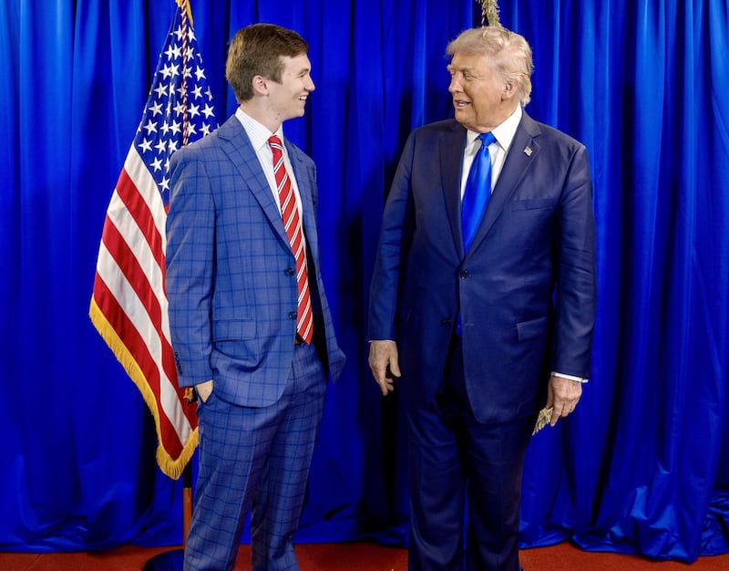 A young man in a checked suit smiling beside Donald Trump in a blue suit. They are standing in front of a U.S. flag