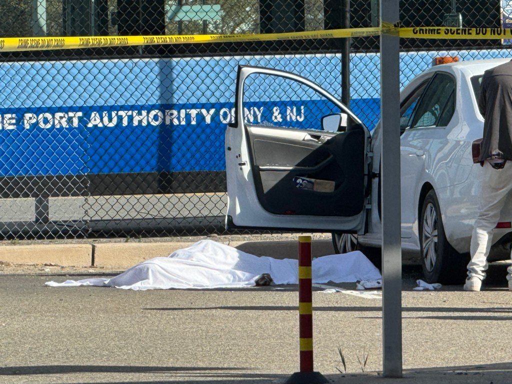 A body covered by a white sheet lies on the asphalt of a parking lot, with a white car door open next to it, behind yellow crime scene tape.