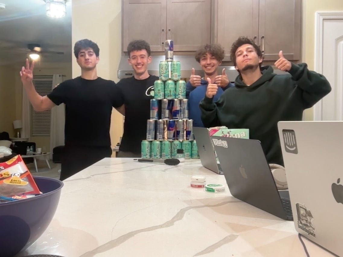 Four young men pose in a kitchen behind a pyramid of stacked energy drink cans, with snacks and laptops on the counter.