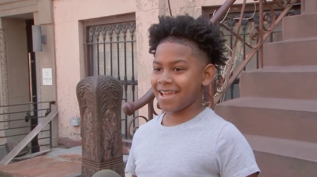 Amir Nur, a 10-year-old boy, smiling while standing in front of a building with stone stairs and an ornate iron railing.