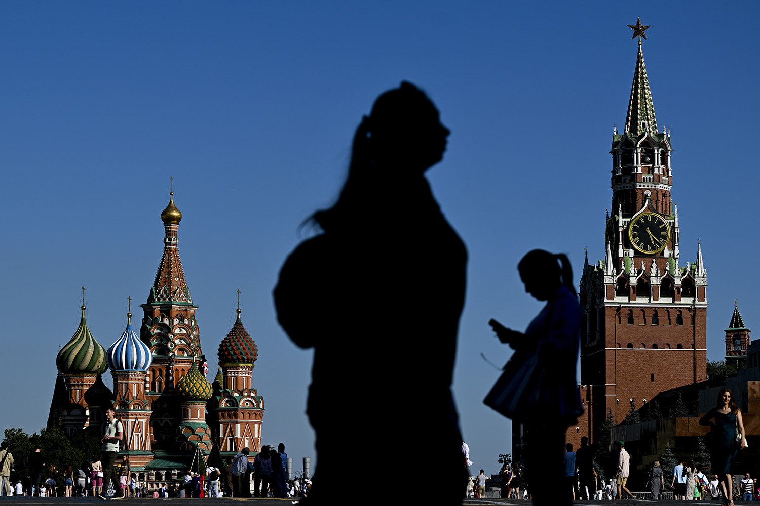 In the foreground is the silhouette of a woman walking in one direction, with the silhouette of another woman, facing the opposite direction and looking at her phone, in the midground. In the background are the towers of the St. Basil Cathedral and the Spasskaya clock tower in the historic Red Square in Moscow, and crowds of people milling through the square.