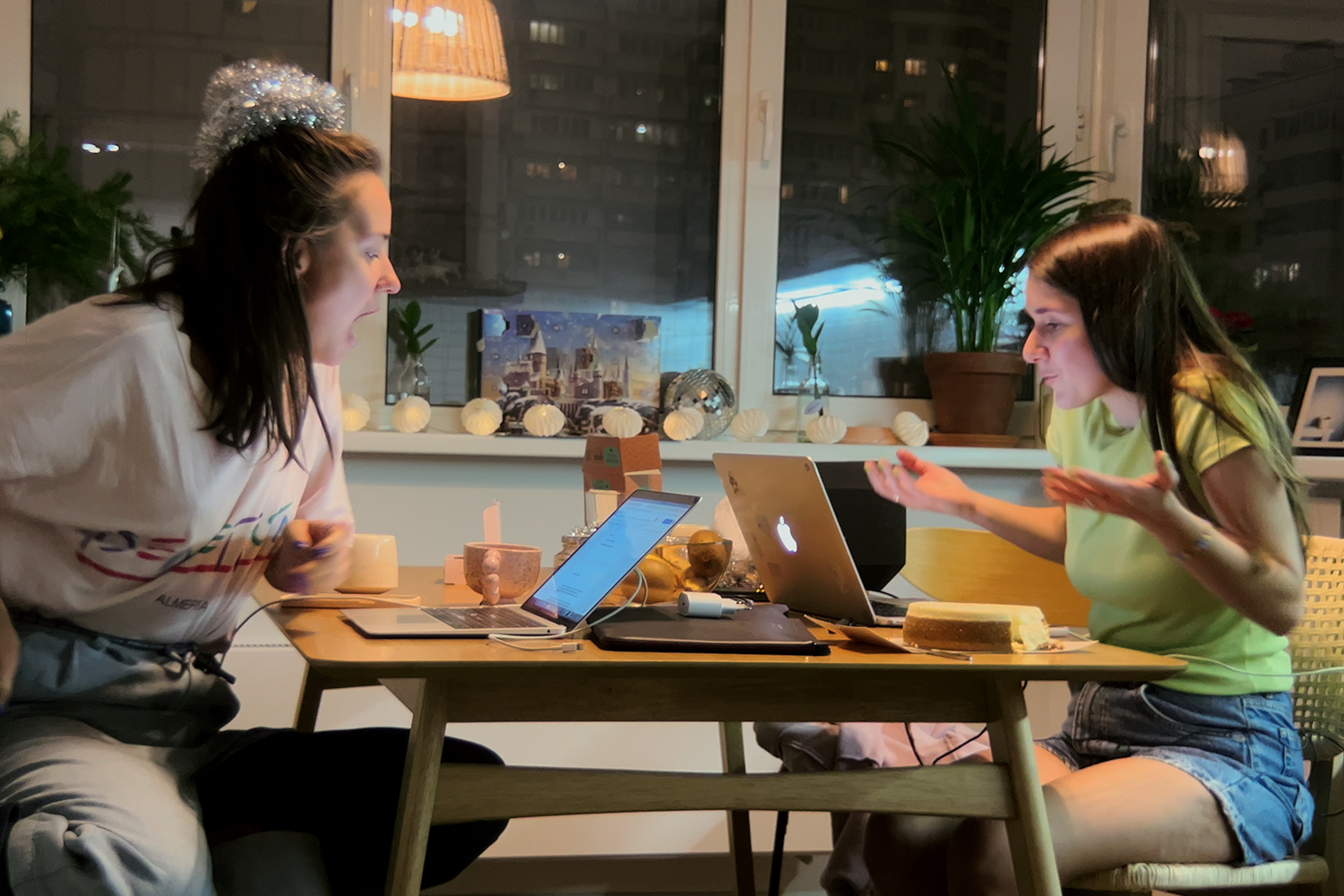 Two women, wearing t-shirts and jeans, sit across from each other at a dining table and look at their laptops. One has her mouth open mid-expression, while the other shrugs with hands raised. Behind them are windows with plants on the sill and apartment buildings visible outside at night.