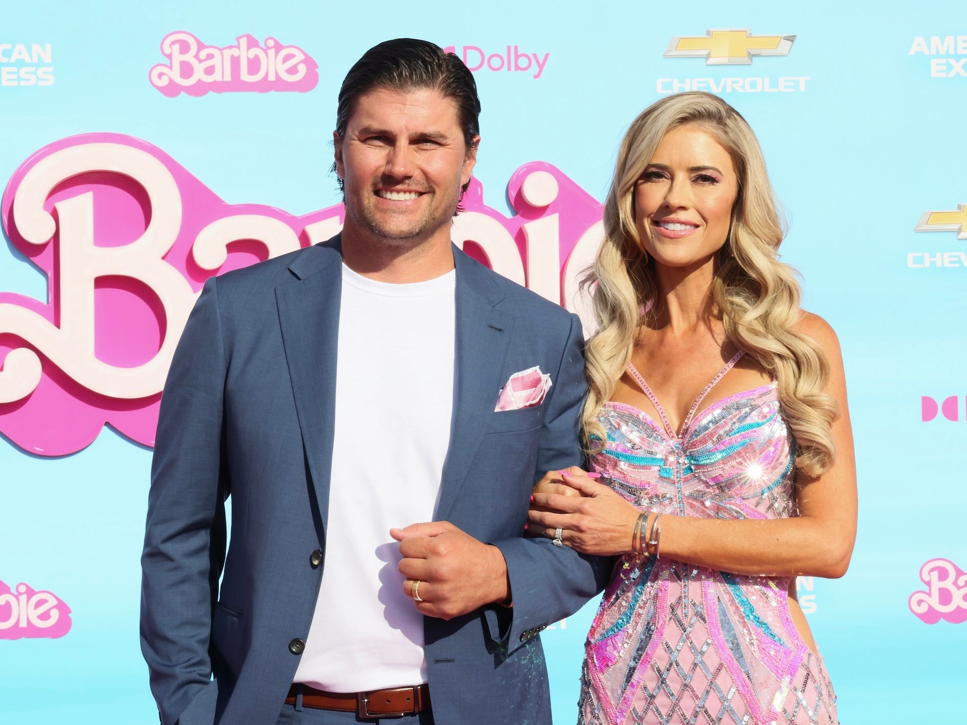 Josh and Christina Hall pose in front of a blue backdrop with the Barbie logo on it.