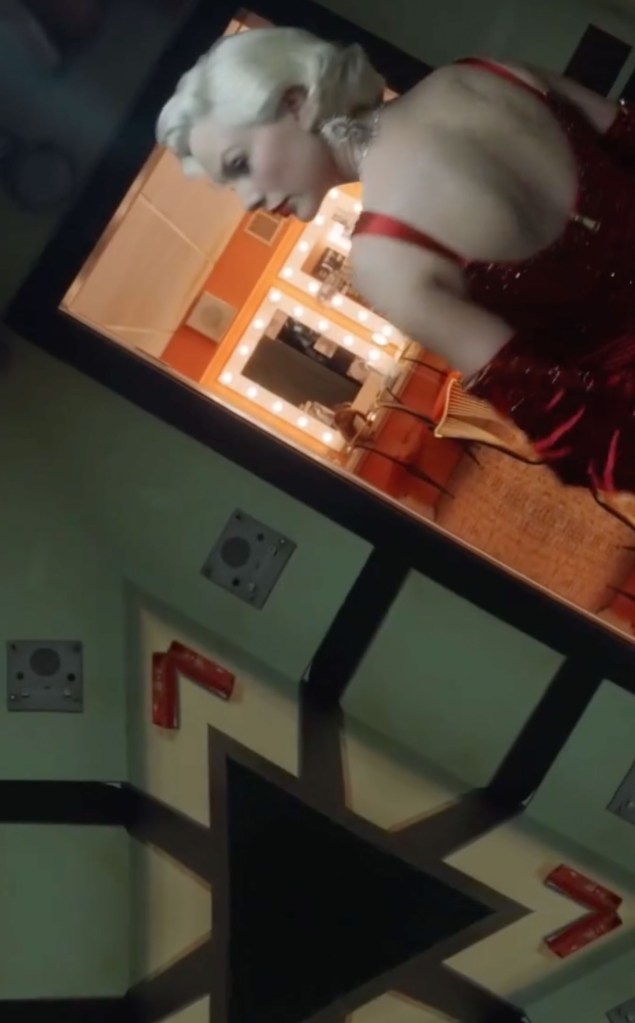 Taylor Swift in a red dress with white blonde hair, looking into a dressing room mirror.