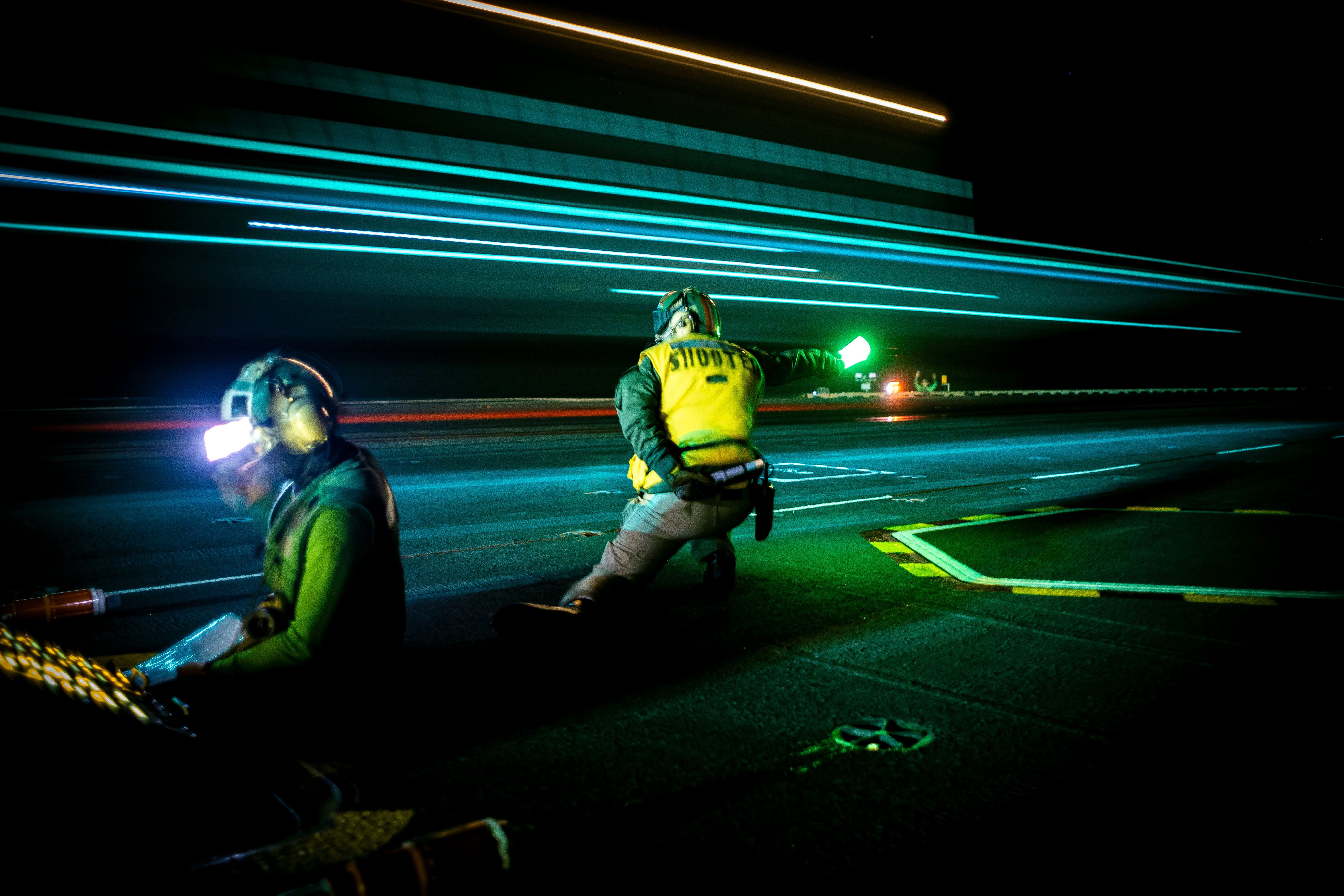 Two aircraft carrier workers are on the top deck as blurring lights of an aircraft soar by them at night.
