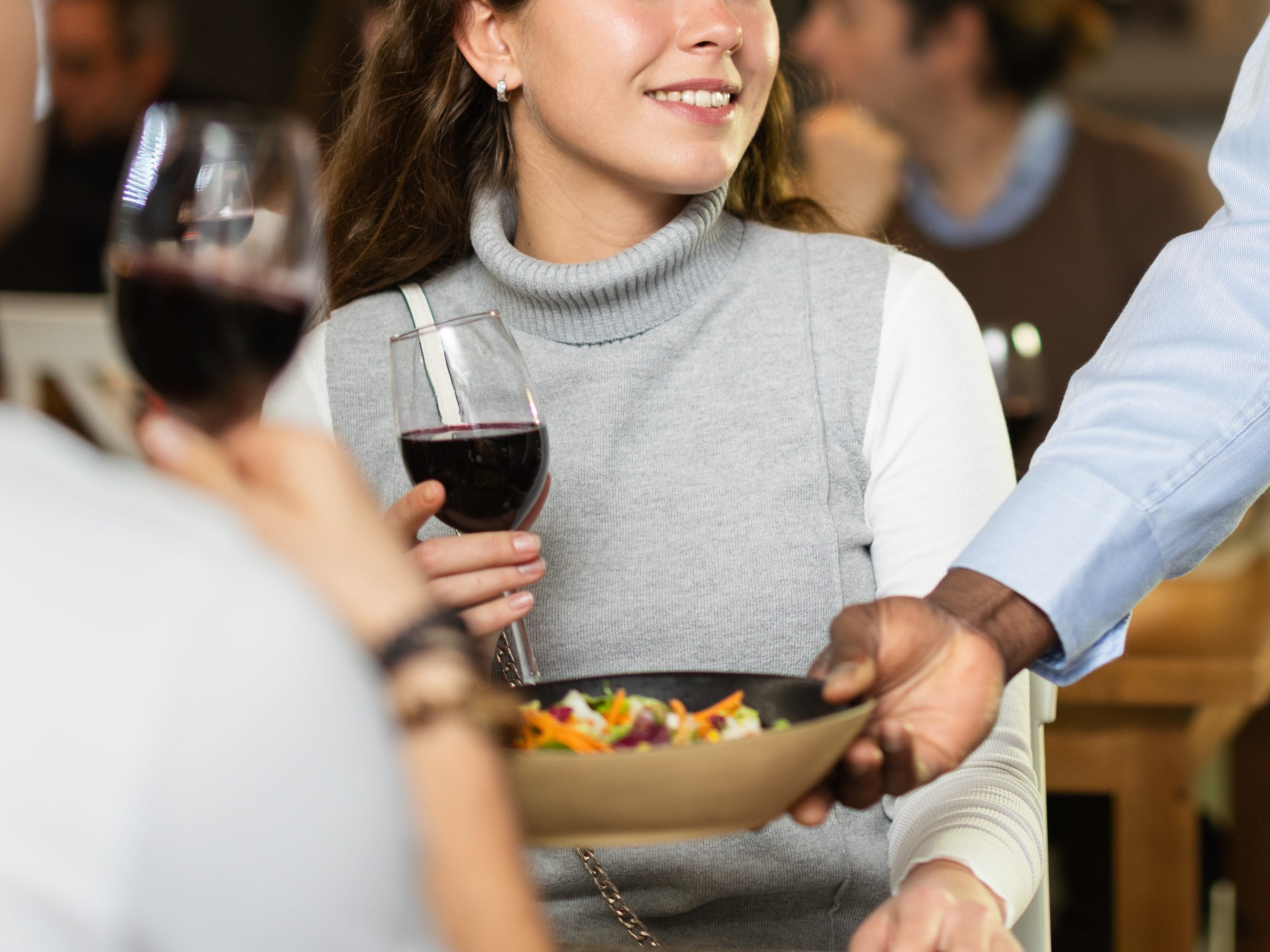 A waiter places a salad in front of a woman holding a glass of red wine.