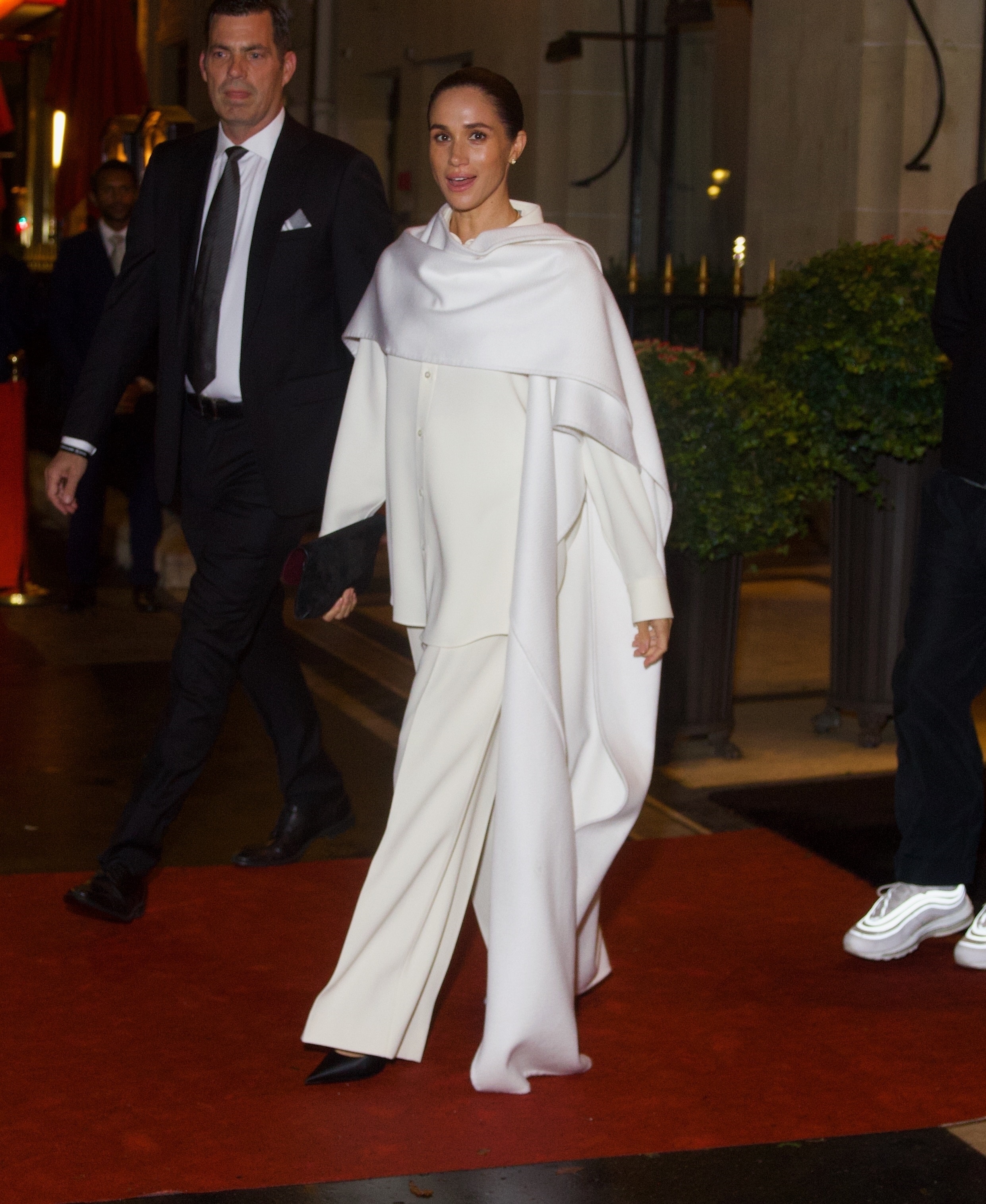 Meghan Markle attending Paris Fashion Week in a white oversized cape over a white button-down shirt, white pants, and black heels.