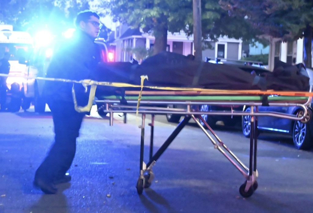 A medical examiner from the NYC Office of the Chief Medical Examiner pushing a stretcher carrying the body of a 37-year-old man, who was fatally shot, on a street in Jamaica, Queens.