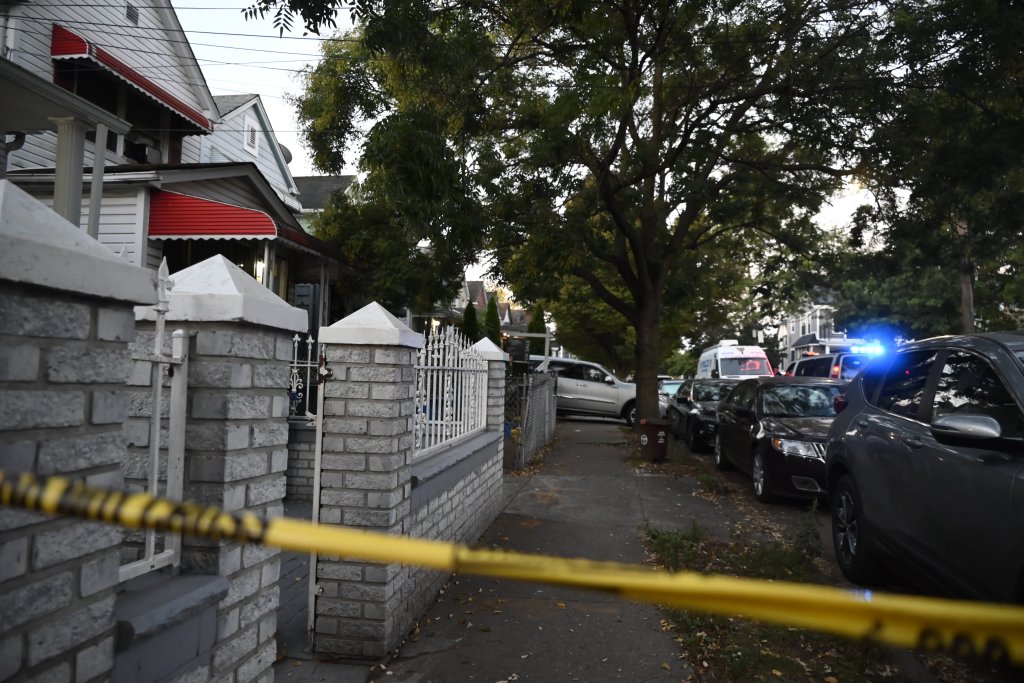 Crime scene tape across the front of a house in Queens, New York, with NYPD officers and vehicles visible in the background.