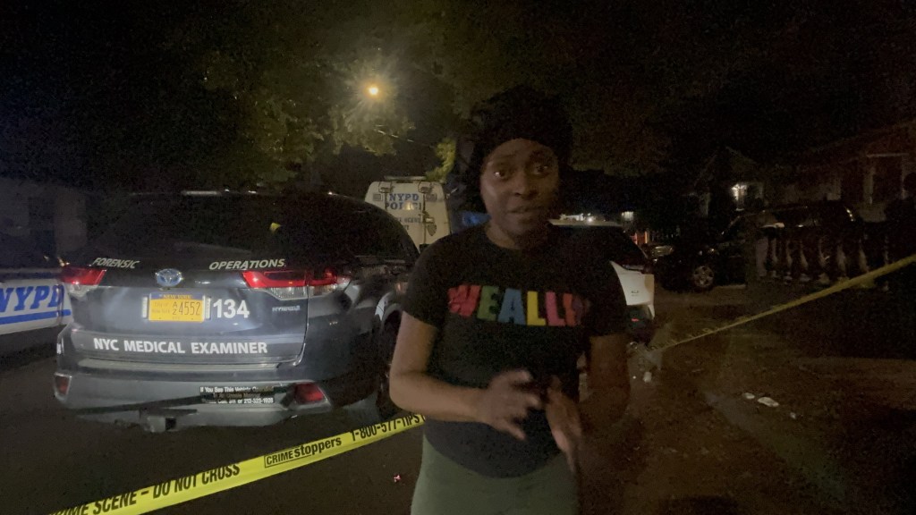 A woman speaks to the press at a crime scene with a NYPD Medical Examiner vehicle in the background.