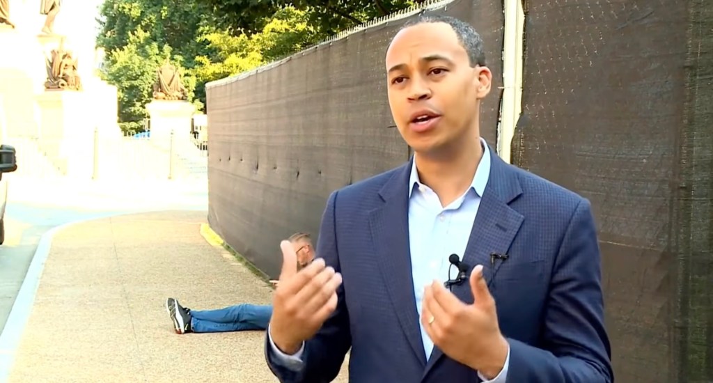 Jay Jones speaking to the camera with a person lying on the ground behind him.