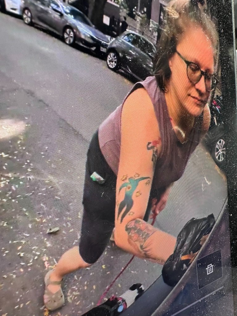 A woman with glasses and tattoos leaning over and holding a black plastic bag against a vehicle, with a 