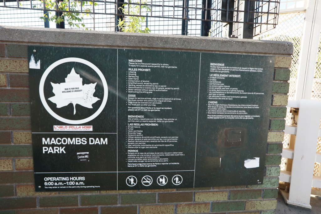 A dark green sign for Macombs Dam Park with rules and operating hours in English, Spanish, and French.