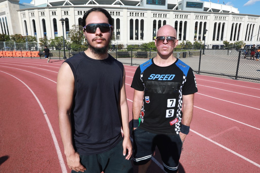 Two men in athletic wear standing on a running track with Yankee Stadium in the background.
