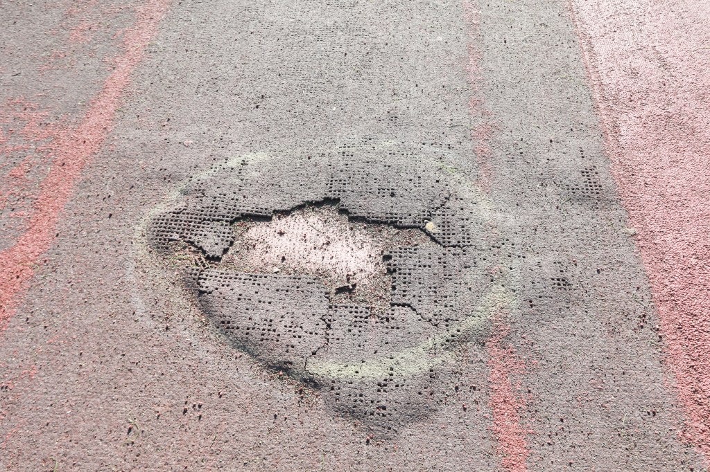 Damaged asphalt surface with exposed grid and red and green markings.