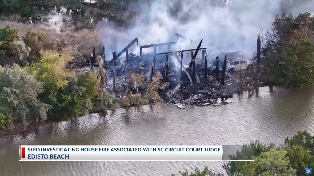 Aerial view of a burned-down house, smoldering with smoke, next to a body of water with green foliage.