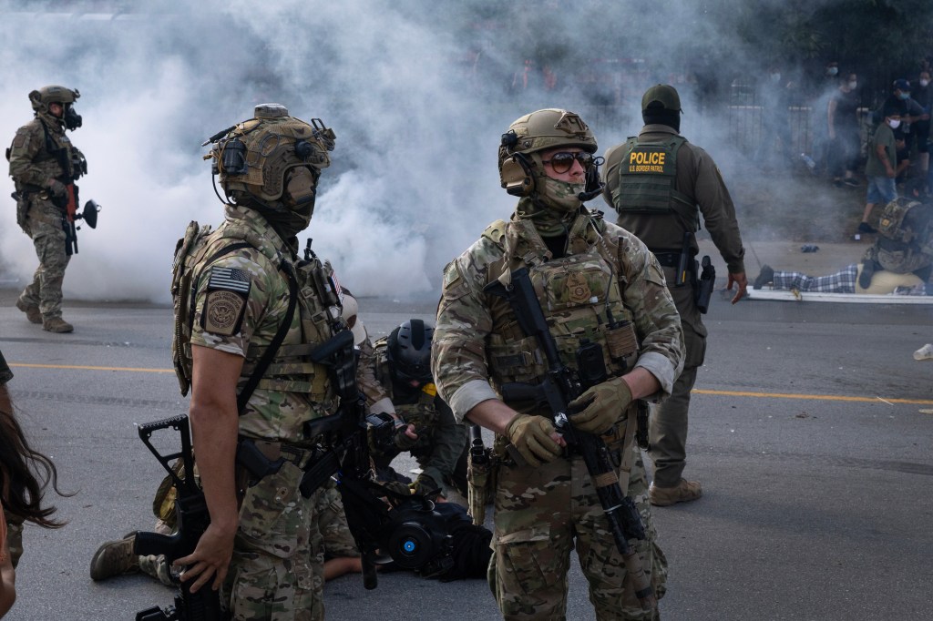 Law enforcement agents in tactical gear standing in tear gas smoke during a confrontation.