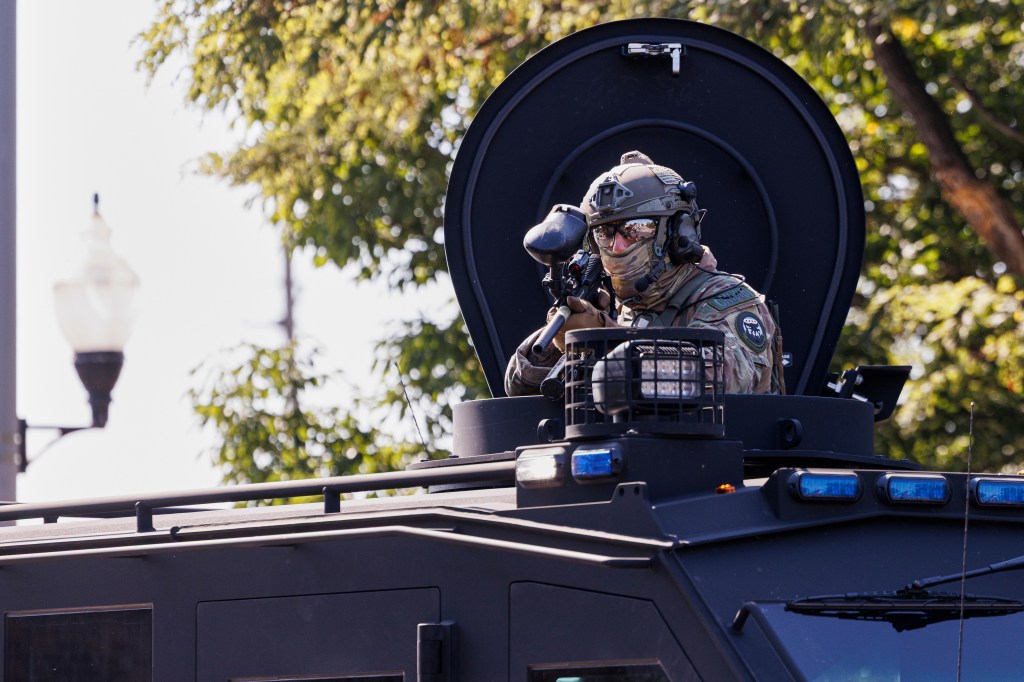A federal officer in tactical gear aims a weapon from the top hatch of an armored vehicle.