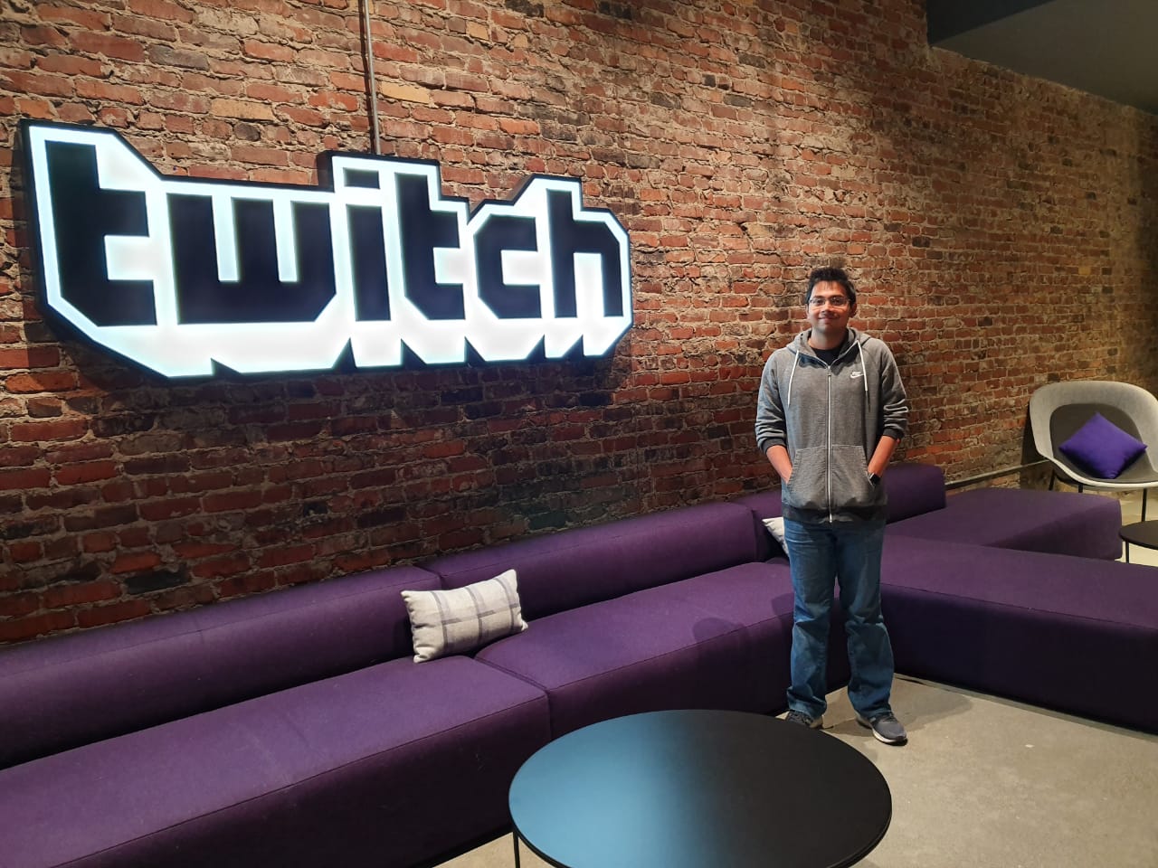 Goutam Nair wears a grey hoodie and smiles in front of the Twitch sign