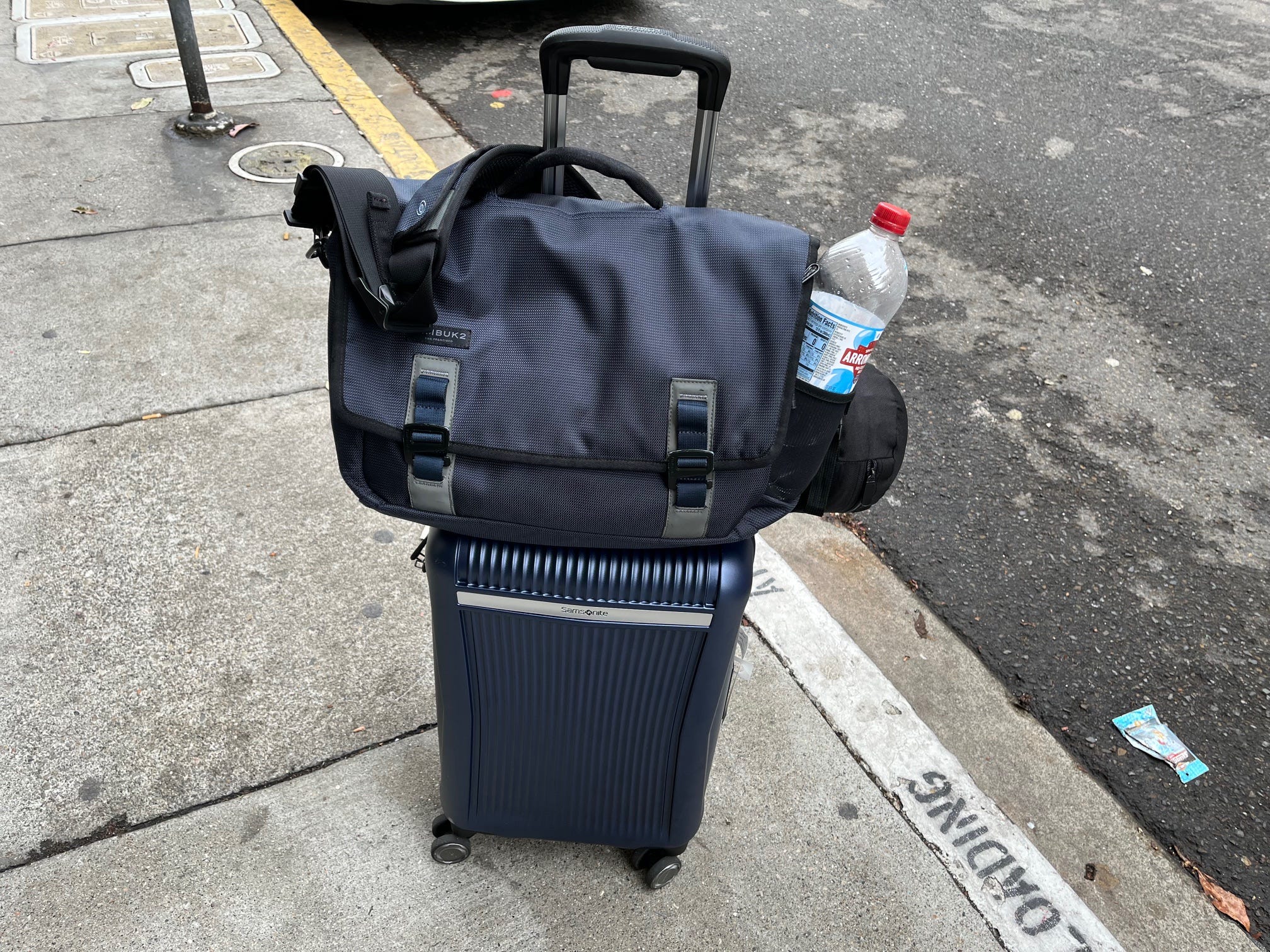 A blue messenger bag with a water bottle sits on top of a blue roller suitcase on a sidewalk