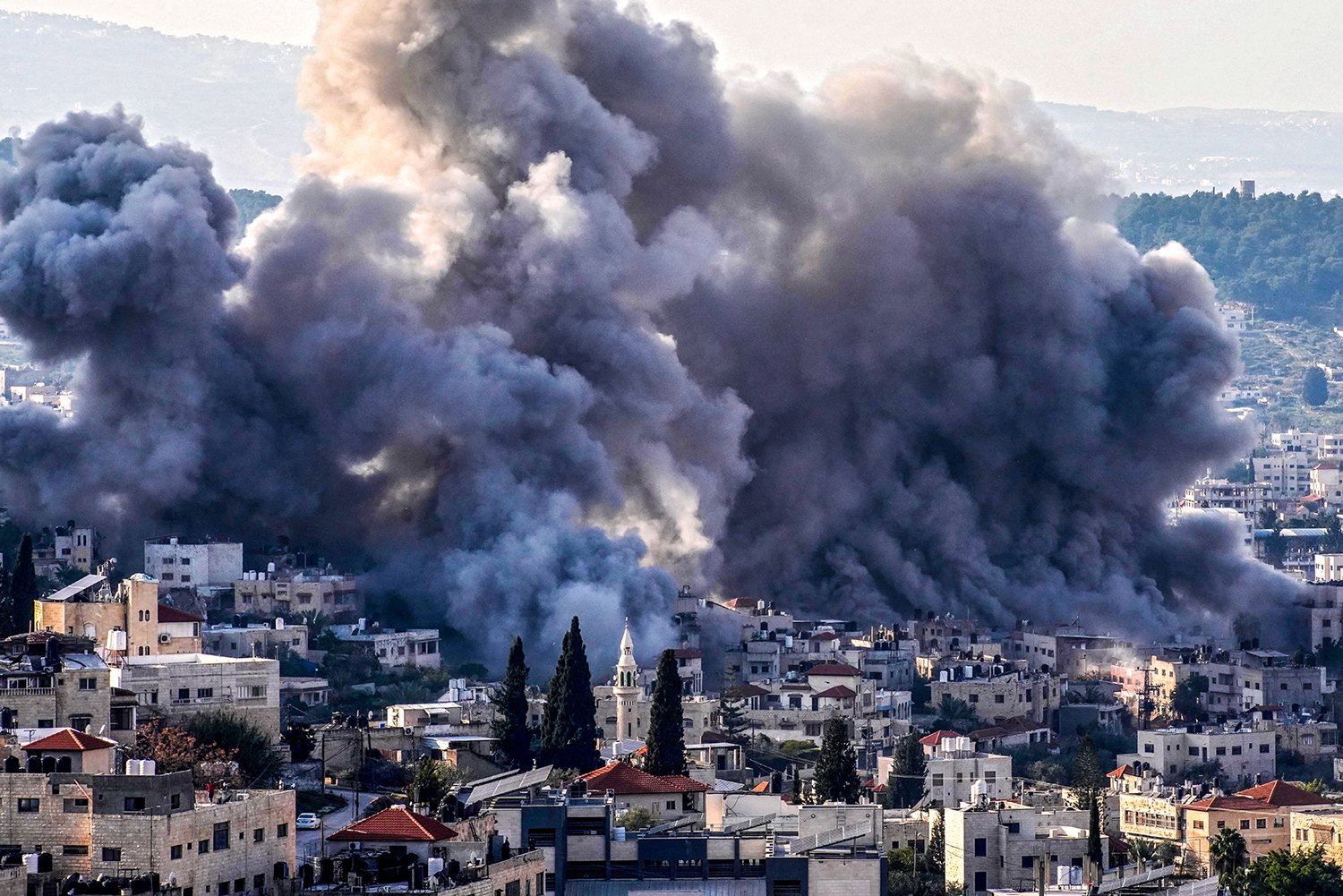 Massive plumes of gray smoke fill the air above densely packed low buildings and houses on a hillside, covering entire city blocks and partly obscuring more tree-covered hills in the distance.