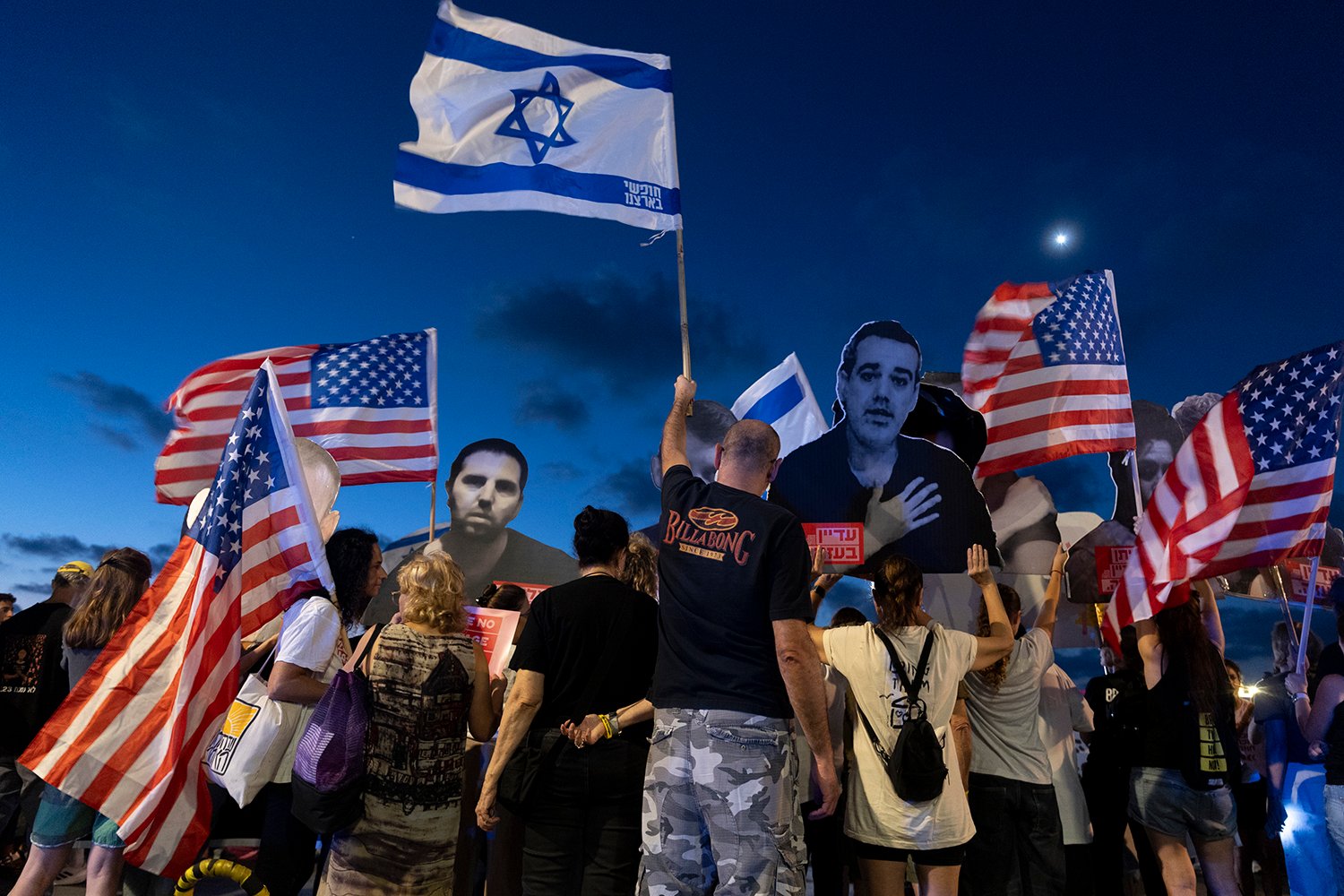 About a dozen people are seen from behind as they hold up sraeli and U.S. flags and photos of hostages beneath a dim sky at dusk.