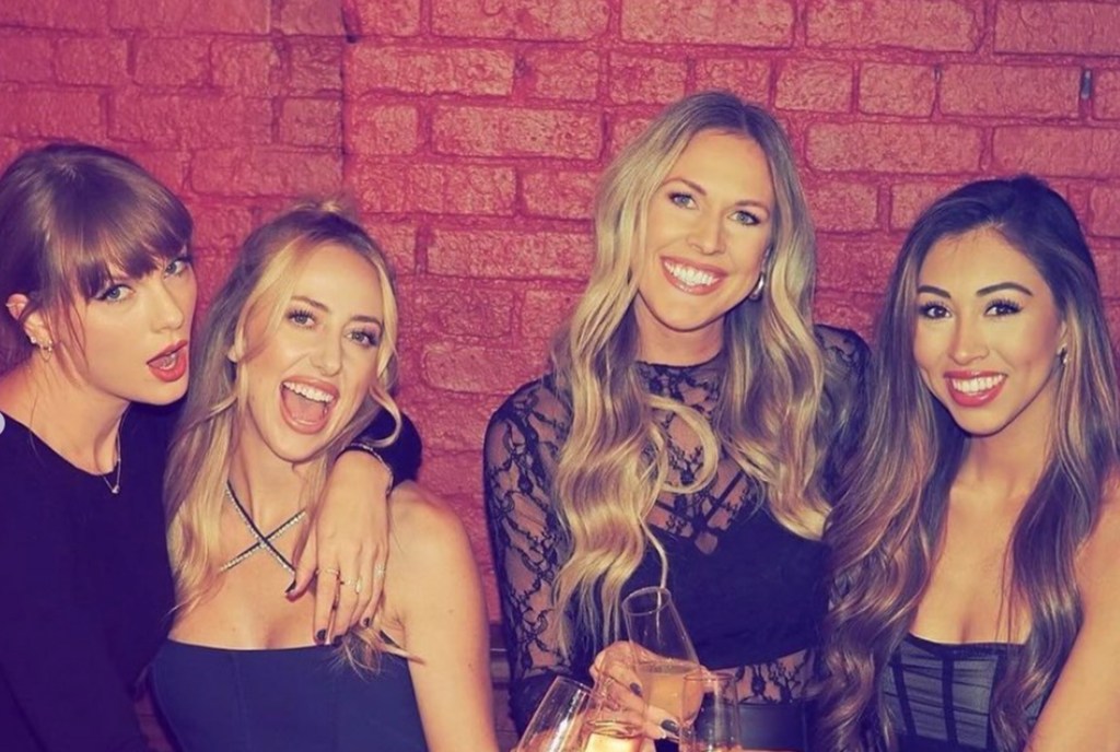 Taylor Swift, Brittany Mahomes, Lyndsay Bell, and Paige Buechele posing together in front of a red brick wall.
