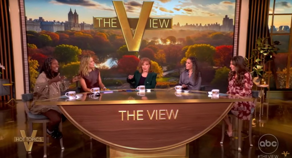 Whoopi Goldberg and fellow hosts on The View set, with a backdrop of autumn trees and the New York City skyline.