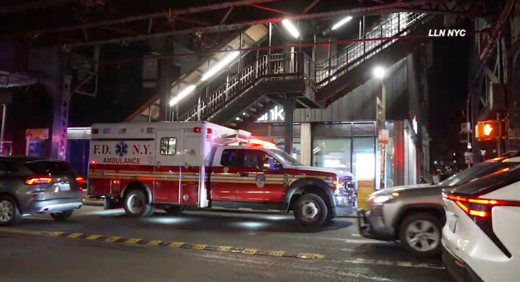 A New York City ambulance parked on a street at night, with an elevated subway station in the background.