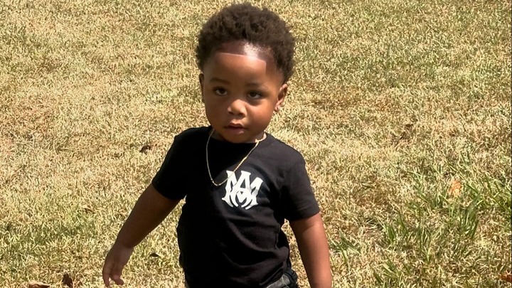 A young boy with a fresh haircut and a gold chain, wearing a black shirt with a white logo, stands in a grassy area.