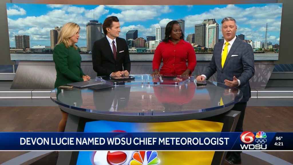 Four news anchors, two men and two women, stand around a glass desk with a city skyline in the background.