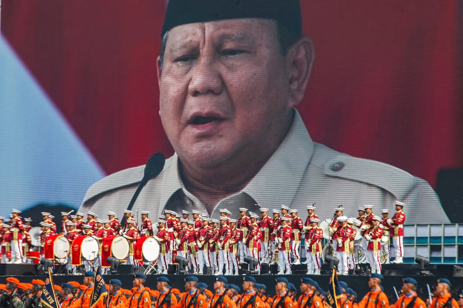 A large screen depicting Prabowo Subianto is behind a band in red and gold jackets on a platform above soldiers dressed in orange.
