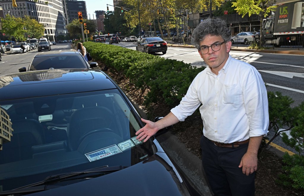 A man in a white shirt and glasses points to multiple parking placards visible on the dashboard of a black car.