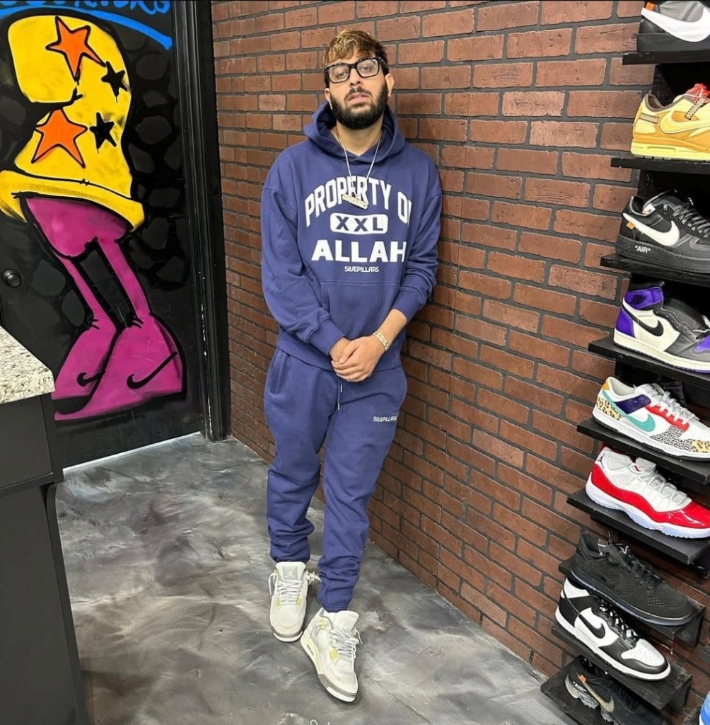 A man wearing a blue tracksuit with “Property of Allah” and sneakers standing in a shoe store.