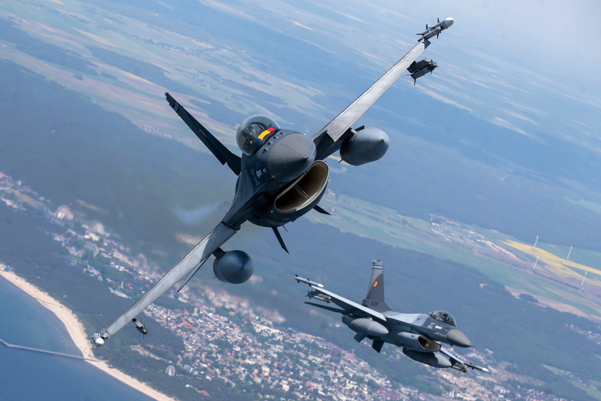 2 grey fighter jets in the sky over some land and some sea, with one banking to its side