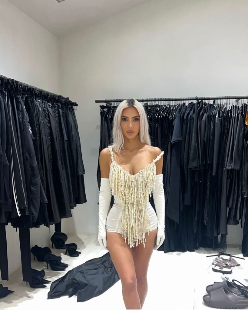 Kim Kardashian in a white pearl-fringed bodysuit and white opera gloves, posing in a room with racks of black clothing.